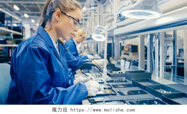 在工厂里工作的女人蓝色工作服和防护眼镜中的女性电子工厂工人正在用螺丝刀组装智能手机。拥有更多员工背景的高科技工厂设施.